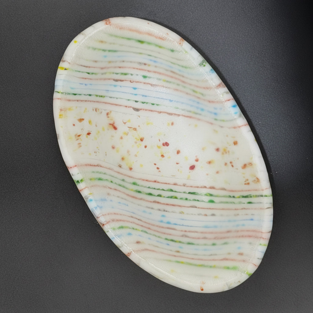 Oval-shaped fused glass dish with multicolored stripes on a dark background