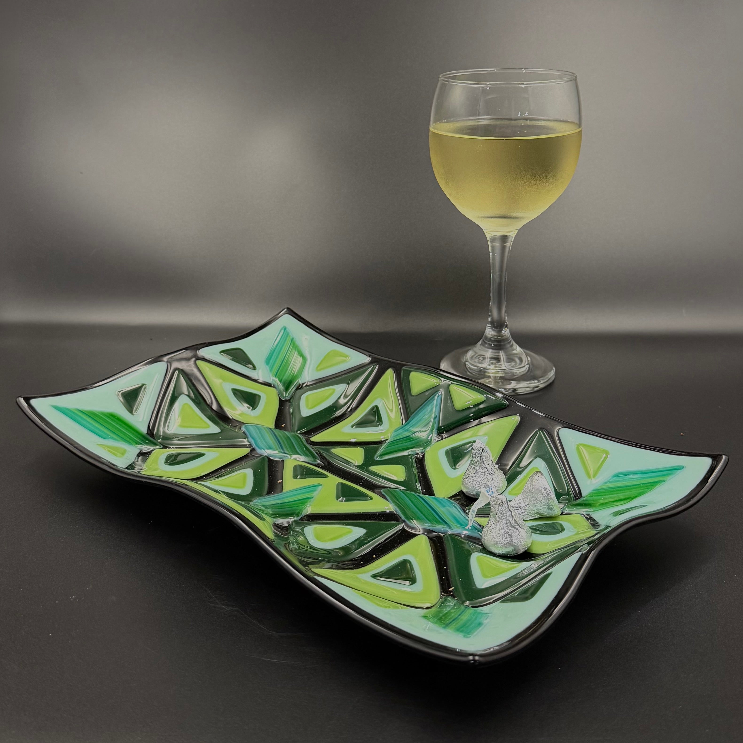 Decorative glass plate with geometric design next to a glass of white wine on a dark surface.